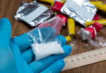 Blue-gloved hand holding bag of white powder, other substances nearby.