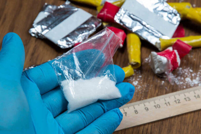 shutterstock_1955608786.jpg featured image Blue-gloved hand holding bag of white powder, other substances nearby.