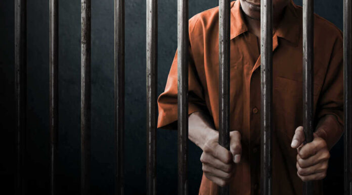 shutterstock_744470836.jpg featured image Man in orange prison uniform behind jail bars.