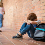 Sad child sitting against brick wall kids running away