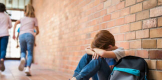 Sad child sitting against brick wall kids running away