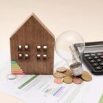 A wooden house model, light bulb, coins, and a calculator on financial documents