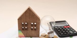 A wooden house model, light bulb, coins, and a calculator on financial documents