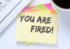 A sticky note with the message 'YOU ARE FIRED' placed on a desk next to a keyboard and a cup of coffee