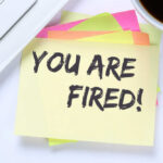 A sticky note with the message 'YOU ARE FIRED' placed on a desk next to a keyboard and a cup of coffee