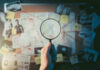 Hand holding magnifying glass over crime investigation evidence board.