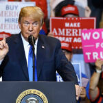 Man speaking at podium with various political signs behind.