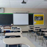 Empty classroom with desks, projector, and chalkboard.