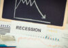 A sign reading 'RECESSION' in front of various financial charts and a downward trend graph