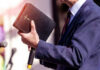 MAGA Pastor’s Run EXPLODES Over Flirty Texts Man holding a Bible during a speech or presentation.