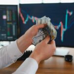 Person holding cash in front of stock market charts on computer screens