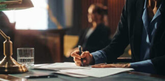 Person writing at desk with documents and glass.