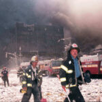 Firefighters walk through debris with smoke and damaged buildings behind them