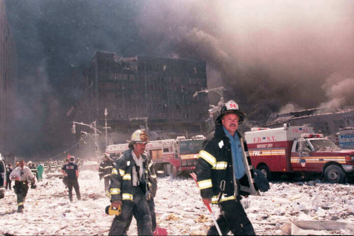 Firefighters walk through debris with smoke and damaged buildings behind them