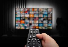 Hand holding a remote control in front of a television displaying various media options